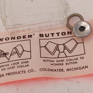 May include: A silver-toned metal Wonder Button with a spring loop, positioned next to a clear plastic container. The item rests on a light pink instruction card featuring black text and diagrams illustrating how to attach the button to a shirt collar.
