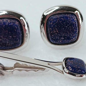 May include: A set of silver-toned cufflinks and a tie clip. The cufflinks and tie clip feature a square, dark blue stone with sparkling inclusions, set within a polished silver frame. The tie clip is a simple, elegant design.
