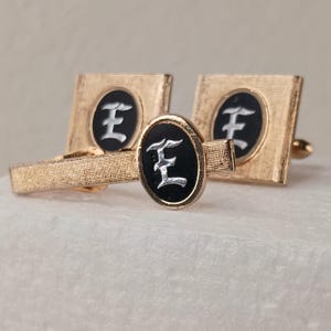 May include: A set of gold-toned metal accessories including two square cufflinks and one tie bar. Each piece features a central oval inlay with a black background and a silver-colored letter E in an old English font, set against a textured gold finish.