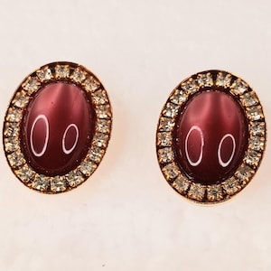 May include: A pair of oval-shaped stud earrings featuring a smooth, deep plum-colored cabochon center stone. Each earring is surrounded by a border of small, clear, sparkling crystals set in a gold-toned metal frame with a polished finish.