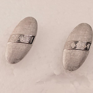 May include: A pair of silver-toned cufflinks with an oval shape and a textured surface. Each cufflink features a decorative band with a repeating geometric pattern. The cufflinks are displayed on a light-colored surface.