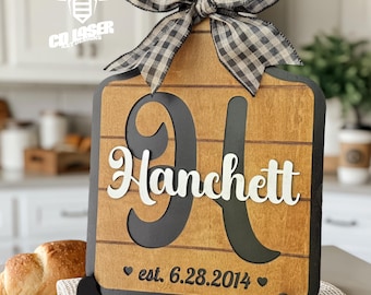 May include: A wooden cutting board sign with a black and white buffalo check bow at the top. The sign features a large black letter H, the white text Hanchett, and the black text est. 6.28.2014 with two small hearts on a light wood background.