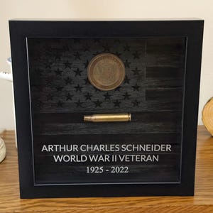 May include: A black shadow box frame containing a dark wood-toned American flag background, a circular United States Army coin, and a horizontal brass-colored bullet casing. The text reads: ARTHUR CHARLES SCHNEIDER WORLD WAR II VETERAN 1925 - 2022.