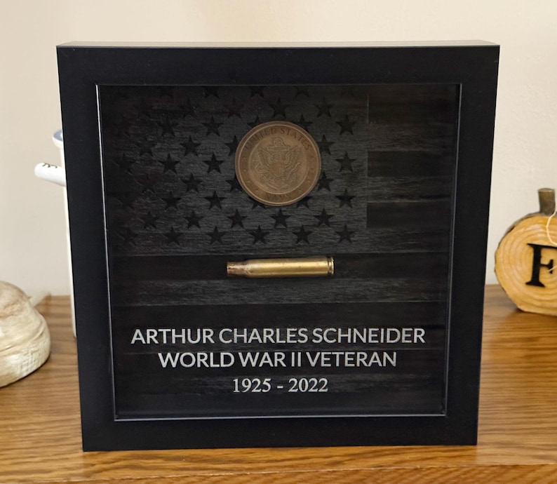 May include: A black shadow box frame containing a dark wood-toned American flag background, a circular United States Army coin, and a horizontal brass-colored bullet casing. The text reads: ARTHUR CHARLES SCHNEIDER WORLD WAR II VETERAN 1925 - 2022.