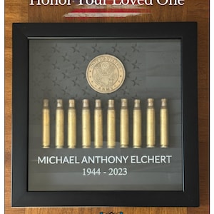 May include: A framed memorial display featuring a U.S. Army coin, brass bullet casings, and the name "MICHAEL ANTHONY ELCHERT 1944-2023" on a gray background with stars. The text "Honor Your Loved One" is above the frame.