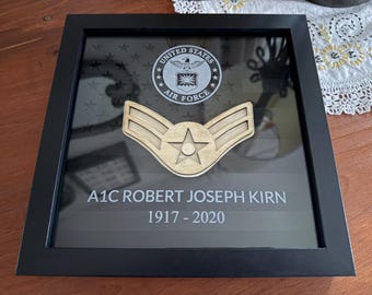 May include: A black square shadow box frame containing a United States Air Force emblem at the top, a wooden Airman First Class rank insignia in the center, and the text A1C ROBERT JOSEPH KIRN 1917 - 2020 printed on the glass surface.