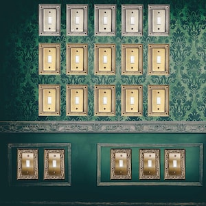 Puede incluir: Una colección de interruptores de luz con marcos dorados y plateados adornados se monta en una pared verde con un patrón de damasco. Una cómoda de madera con una lámpara se encuentra a la derecha. Los interruptores tienen una estética clásica y vintage.