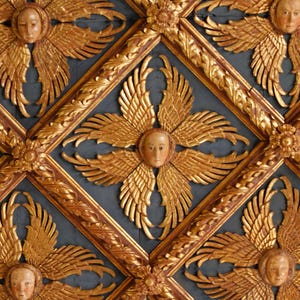 May include: Close-up of a decorative ceiling with a repeating pattern of gold-colored angel faces with wings. The angels are set against a dark blue background, framed by ornate gold trim. The design is symmetrical and detailed.