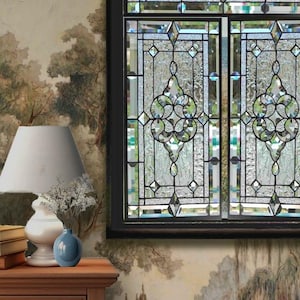 Op de afbeelding: Een glas-in-loodraam met een geometrisch patroon in een zwart frame. Het raam is verdeeld in twee secties, elk met een ander ontwerp. Het glas is helder en heeft een lichte iriserende glans.