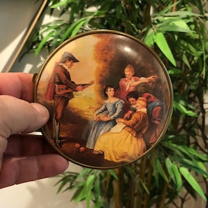 May include: A round, brass-colored trinket box with a painted scene of four people in a garden setting. The scene depicts a man playing a guitar, a woman sitting and reading, and two other women standing. The box has a detailed, vintage style.
