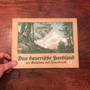 Könnte beinhalten: Ein Vintage-Buchcover mit einer grün-weißen Illustration einer Bergkette und Bäumen. Der Titel des Buches lautet "Das bayerische Hochland mit Salzburg und Innsbruck" mit einer kleinen weißen Blumengrafik auf beiden Seiten des Titels.