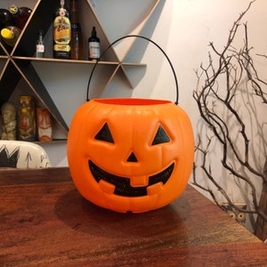 May include: An orange plastic jack-o'-lantern bucket with a black handle. The pumpkin has a carved face with triangular eyes, a triangular nose, and a smiling mouth. A classic Halloween decoration.