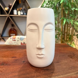 May include: A matte white ceramic vase shaped like a human face with closed eyes, a prominent nose, and defined lips. The cylindrical vessel features a smooth, solid-colored surface and a wide, open top, presenting a neutral and sculptural aesthetic.