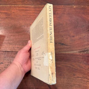 May include: A vintage book titled "French Porcelain" with a tan cover. The spine displays the title in bold black letters. The book's cover has a worn appearance, suggesting age. The book is held at an angle, revealing the side with text and the publisher's name.
