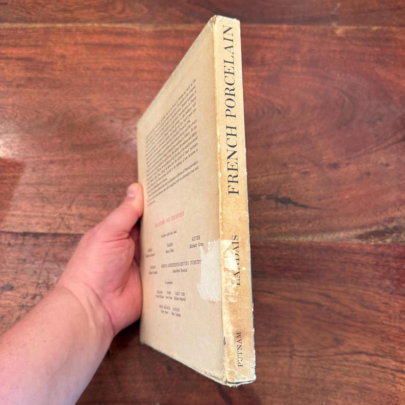 May include: A vintage book titled "French Porcelain" with a tan cover. The spine displays the title in bold black letters. The book's cover has a worn appearance, suggesting age. The book is held at an angle, revealing the side with text and the publisher's name.