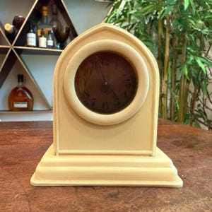 Peut inclure: Horloge de cheminée vintage de couleur ivoire avec un cadran rond. L'horloge a une base rectangulaire et un sommet en forme d'arche. Le cadran comporte des chiffres et des aiguilles noirs. L'horloge est exposée sur une surface en bois.