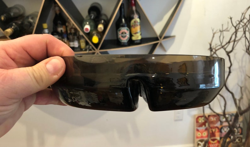 May include: A dark brown glass bowl with a unique, wave-like design. The bowl has a wide, shallow shape and a smooth, glossy finish.