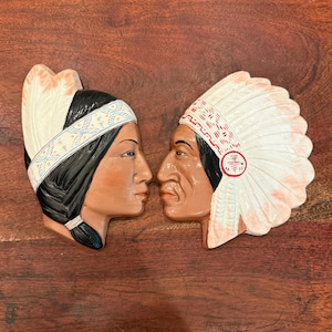 May include: Two ceramic wall plaques depicting the profiles of two individuals facing each other. One figure wears a white headband with blue and orange geometric patterns, while the other wears a large white feathered headdress with red accents.