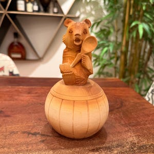 May include: A hand-carved wooden bear figurine holding a spoon, atop a round, lidded container. The bear is detailed with carved fur and facial features. The container has vertical grooves. The wood has a natural, light brown colour.
