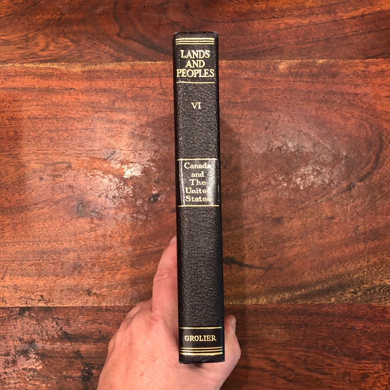 May include: A black leather-bound book titled "Lands and Peoples" volume VI, "Canada and The United States", with gold lettering and the publisher "Grolier" on the spine.