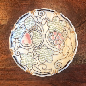 May include: A round ceramic plate with a scalloped edge featuring a hand-painted design of a pink pear and a cluster of pink grapes surrounded by green leaves. The artwork is outlined in dark brown with gold accents on a cream-coloured background.