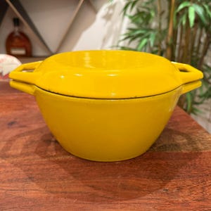 Può includere: Una casseruola olandese in ghisa giallo brillante con coperchio abbinato. La pentola ha due maniglie laterali e poggia su una superficie di legno. Questa pentola è progettata per l'uso sul piano cottura o in forno.