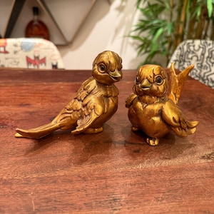 May include: Two gold-coloured bird figurines. One bird is perched with a long tail, the other has a shorter tail. The birds are detailed with feathers and are placed on a wooden surface. Decorative home decor.