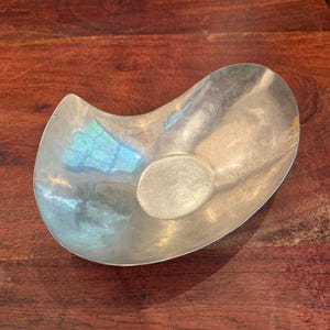 Puede incluir: Un cuenco plateado de forma orgánica con una pequeña hendidura circular. El cuenco tiene una superficie lisa y reflectante, y el interior refleja un toque de azul y verde. El cuenco está sobre una superficie de madera oscura.