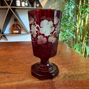 Puede incluir: Una copa de vino de color rojo rubí con base hexagonal y cuenco facetado. La copa presenta un diseño grabado de hojas y vides de uva. La copa mide aproximadamente 18 cm de altura.