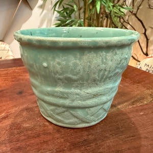 May include: A light turquoise ceramic planter with a wide, flared rim and a textured, basket-weave pattern around the base. The surface features a weathered, matte finish with subtle variations in color and a slightly uneven, rustic texture.