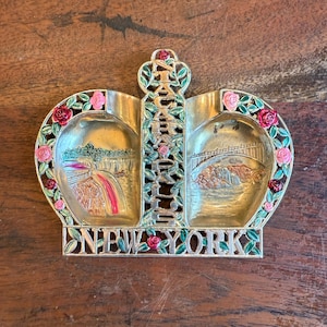 May include: A gold-colored metal souvenir dish shaped like a crown with the words "Niagara Falls New York" around the edge. The dish has two sections, each with a different image of Niagara Falls. One section shows the falls with a rainbow, and the other section shows a bridge over the falls.