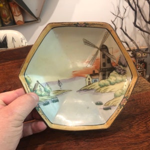 May include: A hexagonal porcelain dish with a hand-painted scene of a windmill, houses, and a sailboat on a body of water. The dish has a gold rim and a white background with a light blue and green color scheme.