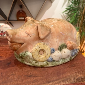 May include: A ceramic pig-shaped serving dish with a glossy peach-colored finish. The pig is depicted with closed eyes and a smiling mouth, surrounded by sculpted details of pineapple slices, blueberries, and mushrooms at its base.