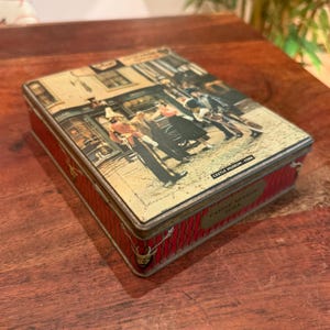 May include: A rectangular metal storage tin featuring a vintage street scene illustration on the lid with the text CASTLE MUSEUM - YORK. The sides are decorated with a red pattern of vertical wavy lines and small deer head icons in gold.