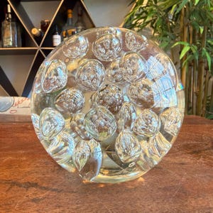 May include: A clear, round glass paperweight with a bubble design. The paperweight is filled with numerous spherical bubbles, creating a textured surface. The glass has a slight yellow tint, and the bubbles reflect light.