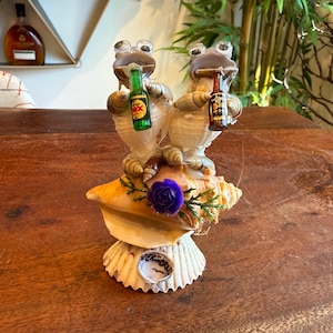 May include: A whimsical seashell sculpture featuring two frog figurines holding miniature beer bottles. The frogs are perched atop a large conch shell, adorned with a purple rose and greenery. The base of the sculpture has a small, round label with text.