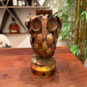 May include: A handcrafted metal owl sculpture with a copper and brass finish. The owl's body is made of overlapping metal pieces, with bottle caps forming the eyes. The base is a circular brass design. A unique decorative item.