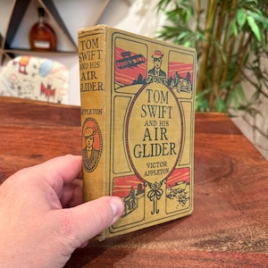 May include: A vintage book titled "Tom Swift and His Air Glider" by Victor Appleton. The cover is yellow with red and black illustrations and text. The book is held in a hand and is on a wooden surface.