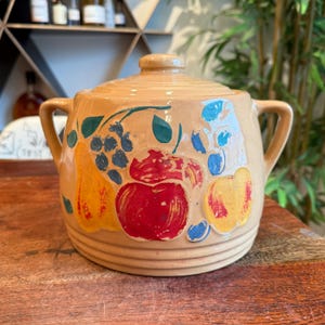 Puede incluir: Un tarro de cerámica vintage con tapa, que presenta un diseño pintado a mano de frutas y hojas en tonos rojos, amarillos, azules y verdes. El tarro tiene dos asas y un cuerpo redondeado.