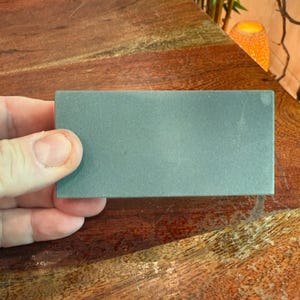 May include: A rectangular, gray sharpening stone is held in a hand. The stone has a slightly textured surface and appears to be made of a dense material. The background is a wooden surface with a warm, reddish-brown tone.