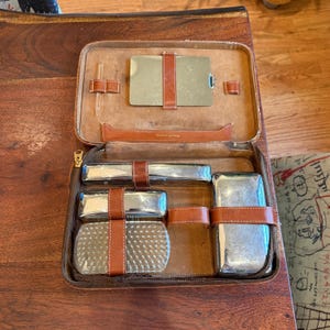 May include: Open brown leather travel grooming kit with a zippered closure. The interior features a mirror, various silver-toned metal containers, and a small glass vial, all secured with brown leather straps.