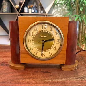May include: Vintage wooden clock with a gold-tone face and black hands. The clock face has the word "United" printed on it. The clock is rectangular with two dark brown cylindrical accents on either side.