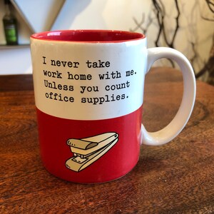 May include: White ceramic mug with a red rim and a red base. The mug features black text that reads "I never take work home with me. Unless you count office supplies." A red and white stapler is depicted on the lower half of the mug.