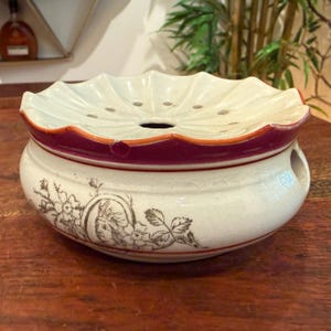 May include: A round ceramic bowl with a scalloped, perforated lid. The base is cream-colored with a dark brown floral and portrait illustration. A wide maroon band encircles the upper rim, which features a small chip. A circular opening is on the side.