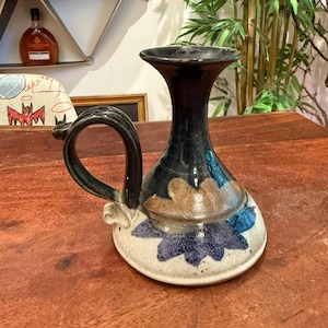 May include: A handcrafted ceramic vase with a dark blue neck and a wide, round base. The vase features a handle and is adorned with brown and blue floral designs on a white background. The vase is a decorative item.
