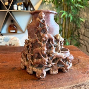 Puede incluir: Jarrón de piedra tallada de color marrón rojizo con un relieve detallado de monos trepando y jugando. El jarrón tiene una base ancha y una parte superior acampanada, con una pequeña abertura lateral. Un artículo decorativo único.