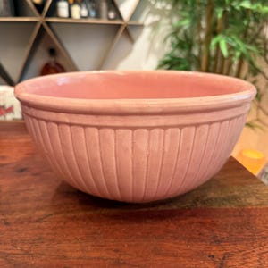 May include: A light pink ceramic mixing bowl with a glossy finish and a rounded rim. The exterior of the bowl features a series of vertical, recessed fluted grooves that extend from the base toward the top edge, creating a textured pattern.
