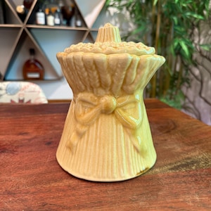 Op de afbeelding: Een keramische pot in de vorm van een bosje tarwe met een strik. De pot is lichtgeel en heeft een deksel.