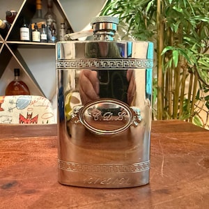 May include: A silver-coloured flask with a screw-on cap and a hinged spout. The flask has a rectangular shape with decorative Greek key borders and an oval emblem with the initials "C.D.L." engraved on the front. The bottom of the flask has the numbers "10120101".