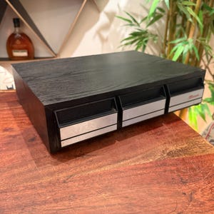 May include: A black desktop storage unit featuring three horizontal drawers with silver-toned front panels. The unit has a wood-grain texture on the top surface and integrated handles on each drawer. The rightmost drawer displays a small red logo.